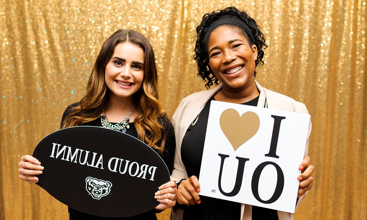 Two women standing together holding signs with text "I heart OU" and "Proud Alumni"